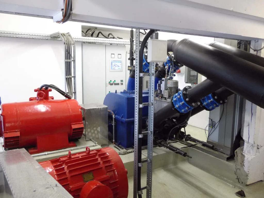 4-düsige Pelton Turbine Gütenbach Heller Maschinenraum mit 4 düsiger Peltonturbine mit schwarzen Rohren, blauen Flansch, roter Generator, montiert auf einem Betonsockel, vorne eine weisse GSM LTE Antenne, hinten eine graue Schaltanlage mit Bildschirm.