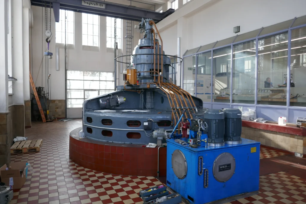 6kV Kaplanturbine mit Statischer Erregung Große Kaplanturbine in einer Maschinenhalle mit Deckenkran, rechts im Bild Blick zum Leitstand, vorne im Bild ein blauer Hydrauliktank mit Pumpen und Sensoren.