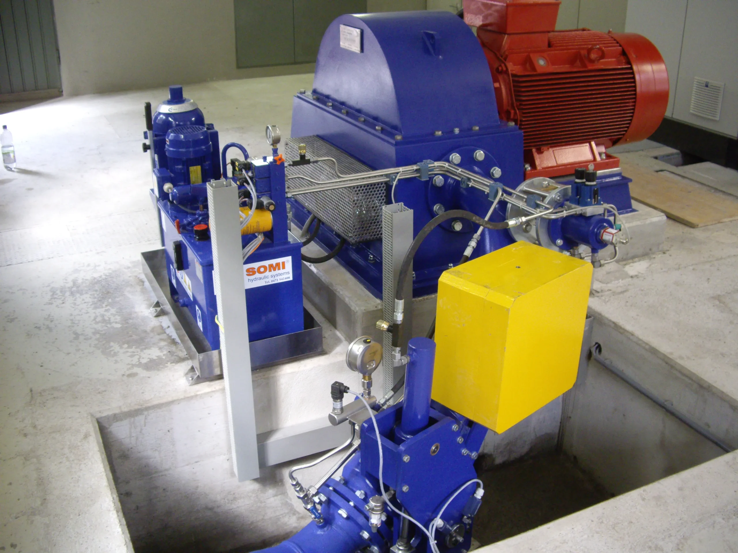 Pelton-Turbine mit Hydrauliksystem und gelbem Absperrschieber in einer technischen Anlage auf Betonboden.