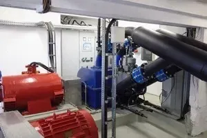 Heller Maschinenraum mit 4 düsiger Peltonturbine mit schwarzen Rohren, blauen Flansch, roter Generator, montiert auf einem Betonsockel, vorne eine weisse GSM LTE Antenne, hinten eine graue Schaltanlage mit Bildschirm.